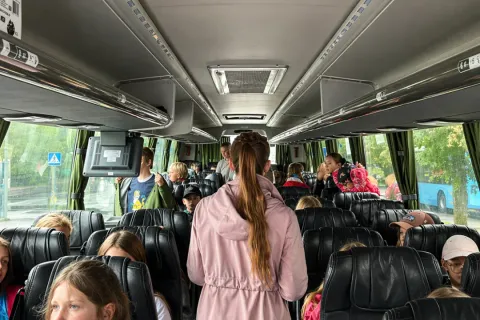 Bussisõidul istuvad lapsed täis istekohtadel, vahekäigus seisab üks täiskasvanu.
